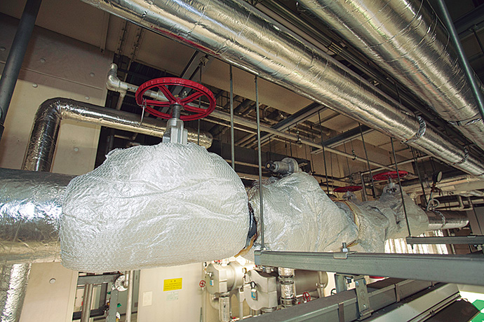 省エネ施策の一例。機械室内の蒸気用配管を断熱ジャケットで包み管内の蒸気の放熱を防いでいる。サーモセンサを使って装着前と装着後の遮熱効果を可視化したところ、機械室内の温度が大幅に下がっていることが明らかとなっている。